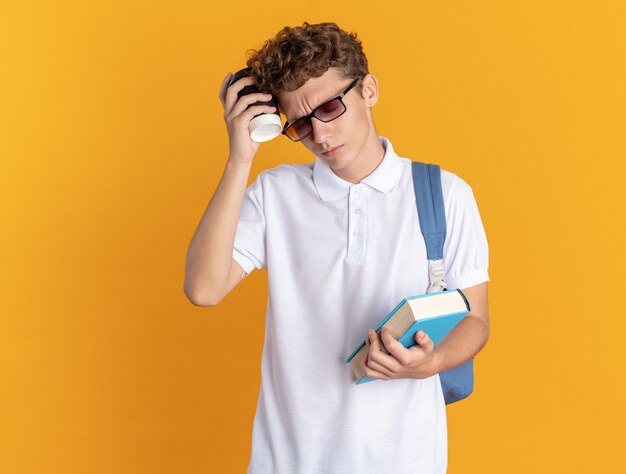 Chico estudiante en ropa casual con gafas con mochila sosteniendo libro y vaso de papel con aspecto cansado