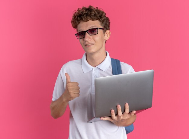 Chico estudiante en ropa casual con gafas con mochila sosteniendo portátil mirando a la cámara sonriendo confiado mostrando Thumbs up