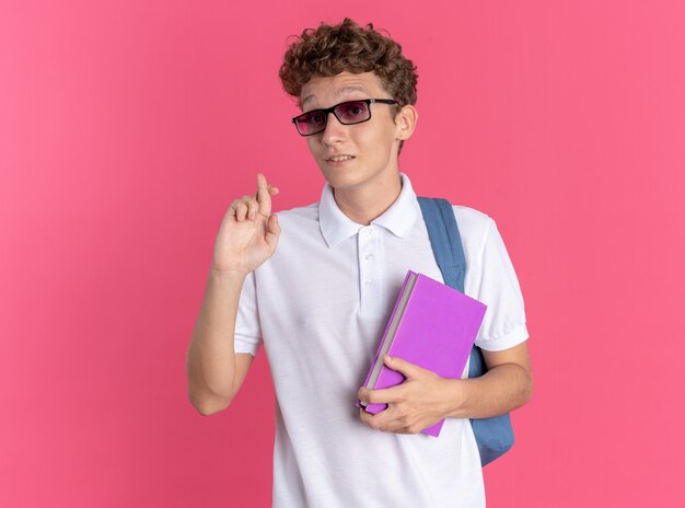 Chico estudiante en ropa casual con gafas con mochila sosteniendo el portátil mirando a la cámara sonriendo haciendo promesa cruzando los dedos