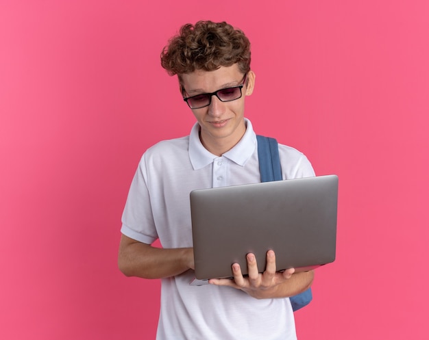 Chico estudiante en ropa casual con gafas con mochila sosteniendo portátil mirándolo con una sonrisa en la cara de pie sobre fondo rosa