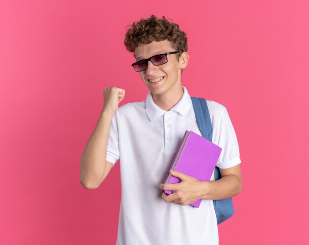 Chico estudiante en ropa casual con gafas con mochila sosteniendo el puño apretado portátil