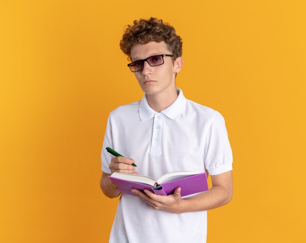 Chico estudiante en ropa casual con gafas sosteniendo libro y lápiz mirando a la cámara