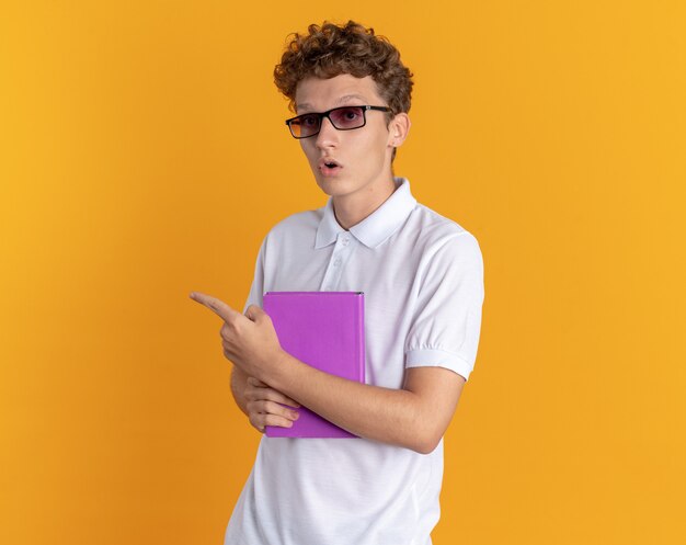 Chico estudiante en ropa casual con gafas sosteniendo el libro mirando a la cámara sorprendido apuntando con el dedo índice hacia el lado de pie sobre fondo naranja