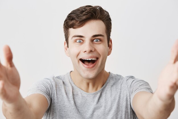 Un chico europeo guapo y feliz sonríe feliz mientras recibe palabras agradables de los padres, muestra dientes blancos y perfectos y estira los brazos hacia la cámara. Joven estudiante masculino se regocija día exitoso