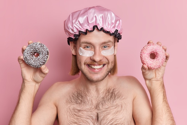 Chico europeo positivo con expresión alegre aplica parches de colágeno debajo de los ojos sostiene donuts dulces usa gorro de ducha está medio desnudo