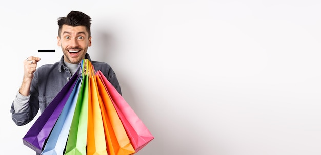 Foto gratuita chico feliz adicto a las compras sosteniendo coloridas bolsas de compras y mostrando una tarjeta de crédito de plástico sonriendo emocionado