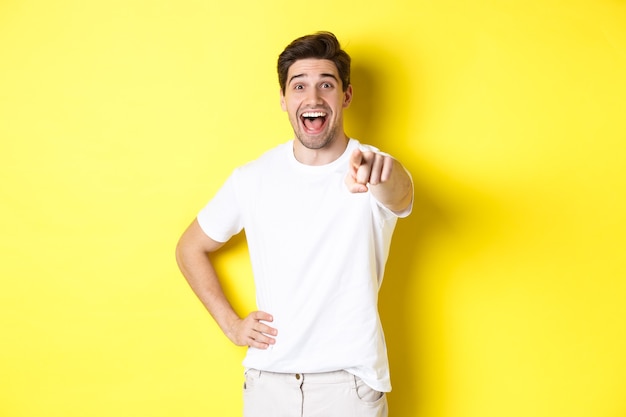 Chico feliz apuntando con el dedo a la cámara y riendo echa un vistazo a algo parado sobre fondo amarillo