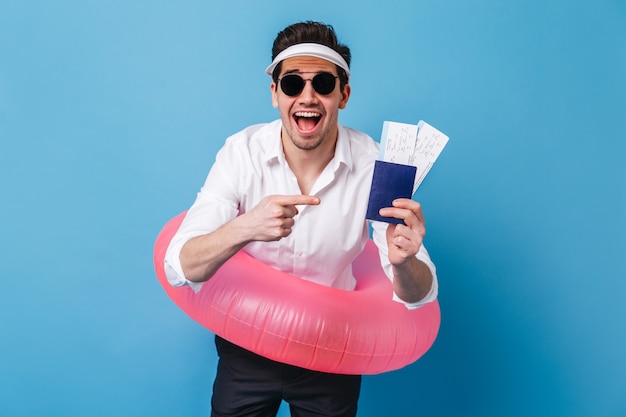 Chico feliz con camisa blanca y pantalón oscuro posa con círculo inflable, documentos y boletos. Retrato de hombre con gafas y gorra en espacio azul.