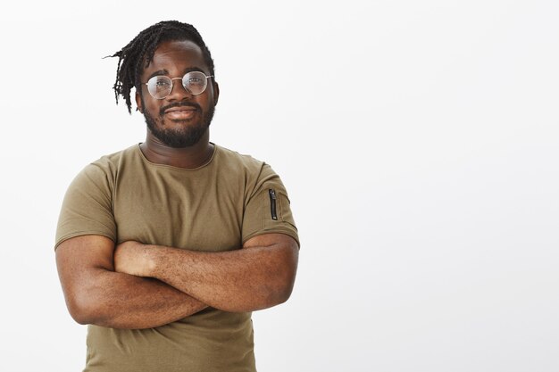 Chico feliz inteligente con gafas y vistiendo una camiseta marrón posando contra la pared blanca