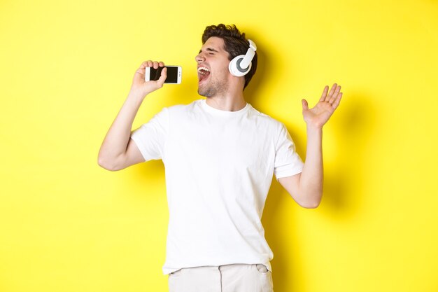 Chico feliz jugando aplicación de karaoke en auriculares, cantando en el micrófono del teléfono inteligente, de pie sobre fondo amarillo.