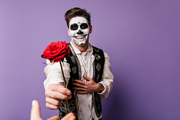 El chico feliz con mirada alegre está agradecido y le da una rosa a su amada. Retrato de interior de hombre con maquillaje de halloween.