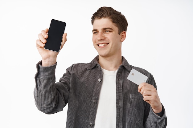 Chico feliz y satisfecho que muestra la tarjeta de crédito, levanta la mano con el teléfono inteligente, muestra la pantalla del teléfono móvil, la interfaz de la aplicación, sonríe complacido, fondo blanco