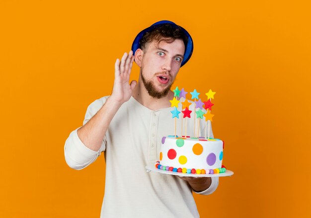 Chico de fiesta eslavo guapo joven curioso con sombrero de fiesta sosteniendo pastel de cumpleaños con estrellas mirando a cámara haciendo no puedo oírle gesto aislado sobre fondo naranja con espacio de copia