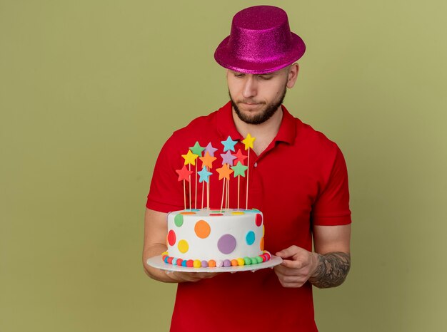 Chico de fiesta eslavo guapo joven pensativo con sombrero de fiesta sosteniendo y mirando el pastel de cumpleaños aislado sobre fondo verde oliva con espacio de copia