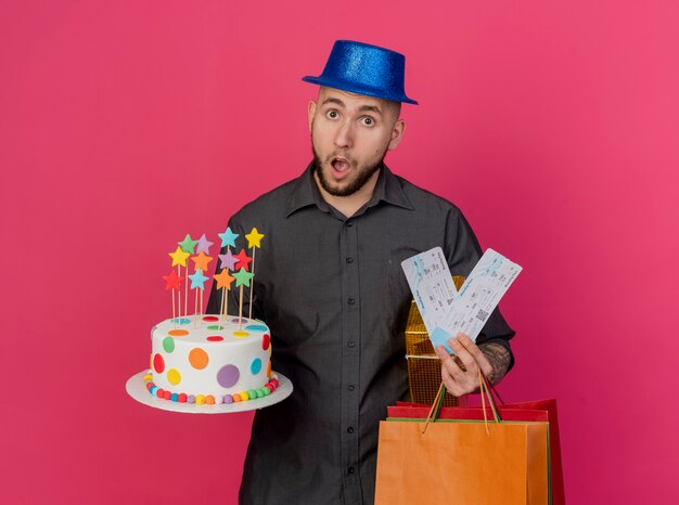 Chico de fiesta eslavo guapo joven sorprendido con sombrero de fiesta con paquete de regalo de dinero de pastel de cumpleaños y bolsas de papel mirando a cámara aislada sobre fondo carmesí