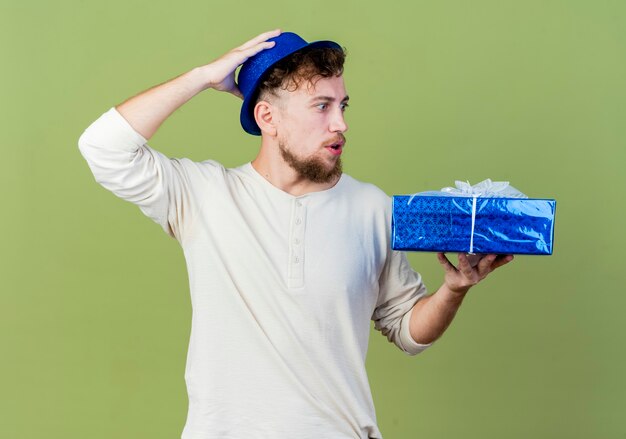 Chico de fiesta eslavo guapo joven sorprendido con sombrero de fiesta sosteniendo y mirando la caja de regalo manteniendo la mano en la cabeza aislada sobre fondo verde oliva