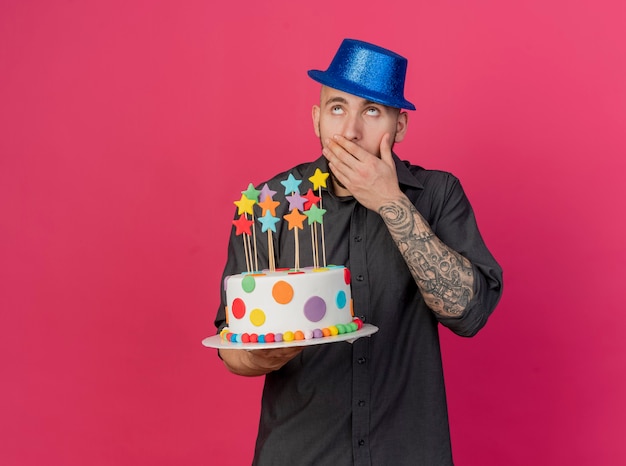 Foto gratuita chico de fiesta eslavo guapo joven sorprendido con sombrero de fiesta sosteniendo pastel de cumpleaños con estrellas mirando hacia arriba manteniendo la mano en la boca aislada sobre fondo carmesí con espacio de copia