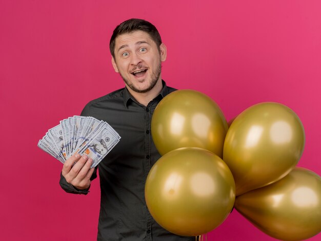 Chico de fiesta joven alegre con camisa negra con dinero en efectivo con globos aislados en rosa