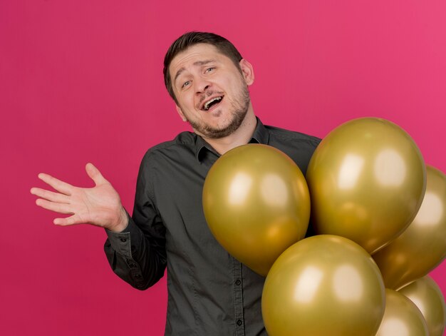 Chico de fiesta joven alegre con camisa negra de pie junto a globos extendiendo la mano aislada en rosa