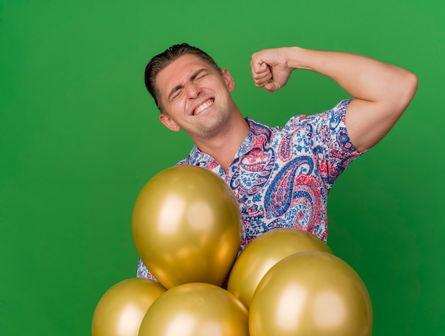 Chico de fiesta joven alegre con los ojos cerrados con camisa colorida de pie detrás de globos mostrando gesto sí aislado en verde