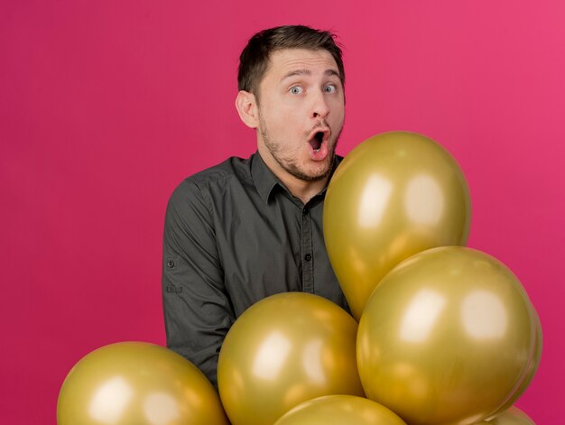 Chico de fiesta joven asustado con camisa negra de pie detrás de globos aislados en rosa