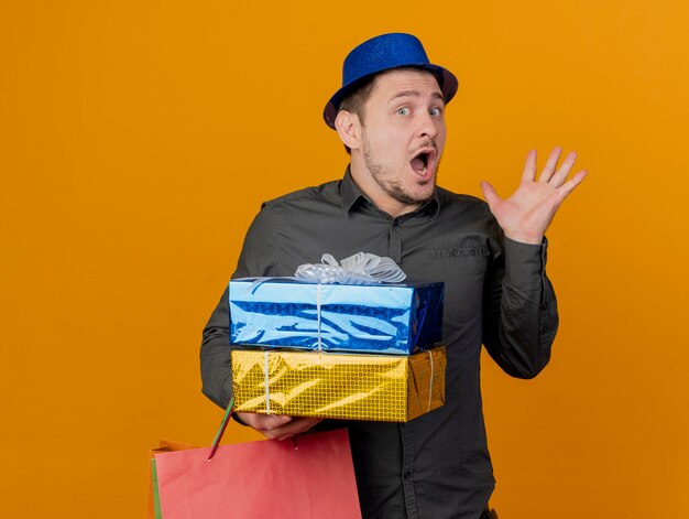 Chico de fiesta joven asustado con sombrero azul sosteniendo cajas de regalo con bolsa extendiendo la mano aislada en naranja