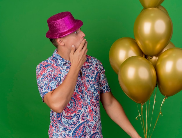 Chico de fiesta joven asustado con sombrero rosa sosteniendo y mirando globos cubiertos la boca con la mano aislada sobre fondo verde