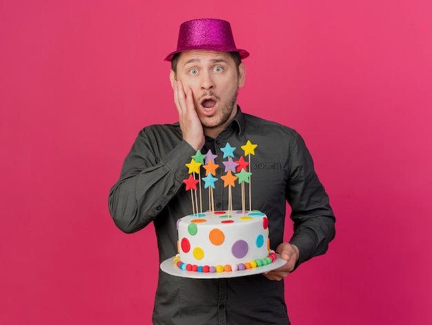 Chico de fiesta joven asustado con sombrero rosa sosteniendo pastel poniendo la mano en la mejilla aislada en rosa