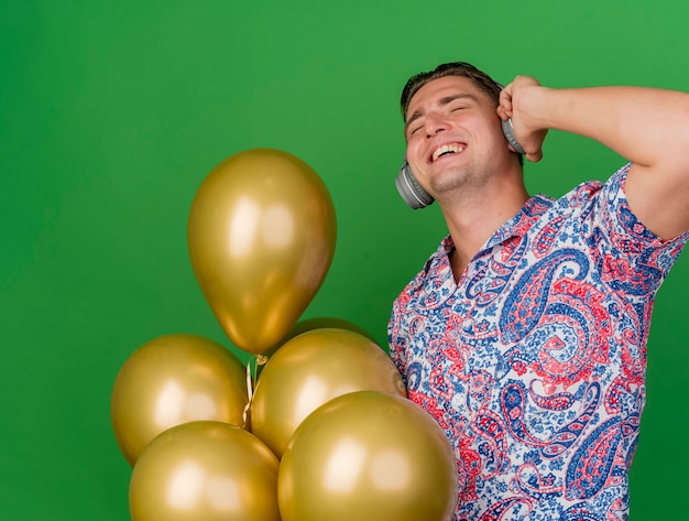 Chico de fiesta joven complacido con los ojos cerrados con camisa colorida y auriculares sosteniendo globos aislados sobre fondo verde