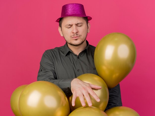 Chico de fiesta joven complacido con los ojos cerrados con sombrero rosa de pie entre globos aislados en rosa