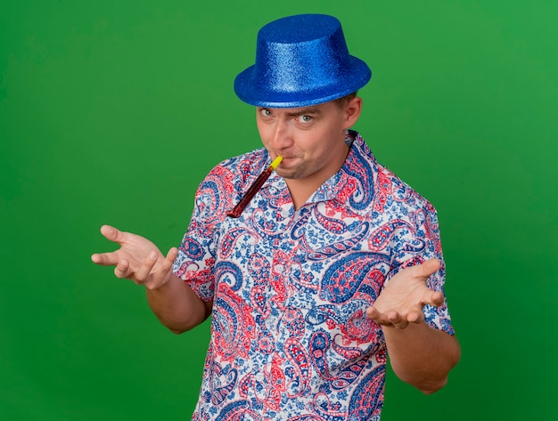 Chico de fiesta joven complacido con sombrero azul soplando soplador de fiesta y extendiendo las manos a la cámara aislada sobre fondo verde