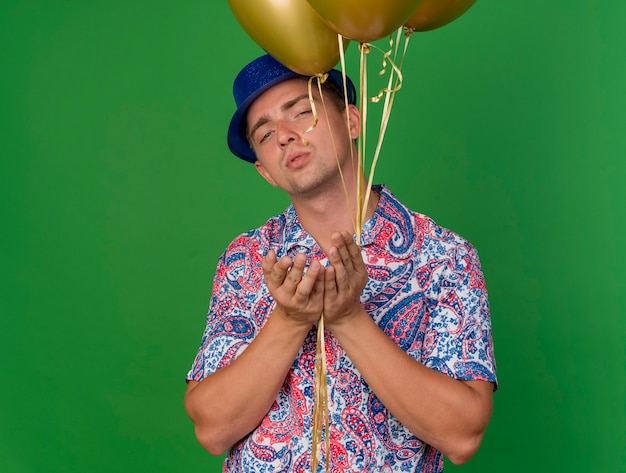 Chico de fiesta joven complacido con sombrero azul sosteniendo globos y mostrando gesto de beso aislado en verde