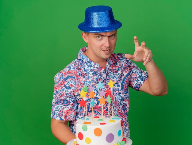 Chico de fiesta joven complacido con sombrero azul sosteniendo pastel mostrando gesto de estilo tigre aislado en verde