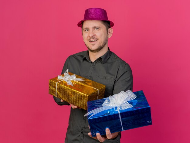 Chico de fiesta joven complacido con sombrero rosa con cajas de regalo aisladas en rosa