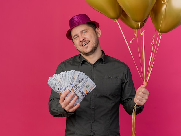 Chico de fiesta joven complacido con sombrero rosa con dinero en efectivo con globos aislados en rosa