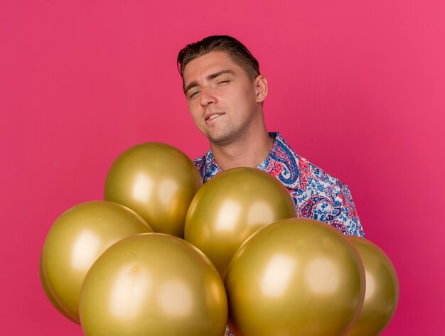 Chico de fiesta joven complacido con sombrero rosa de pie entre globos aislados en rosa