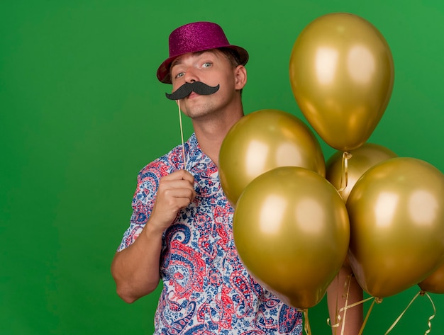 Chico de fiesta joven confiado con sombrero rosa sosteniendo globos poniendo bigote falso en palo aislado en verde