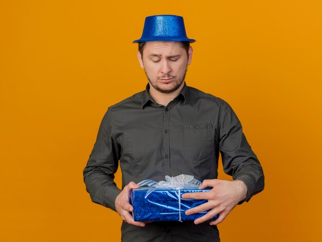 Chico de fiesta joven confundido con sombrero azul sosteniendo y mirando la caja de regalo aislada en naranja