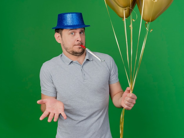 Chico de fiesta joven confundido con sombrero de fiesta soplando silbato sosteniendo globos y extendiendo la mano aislada en verde