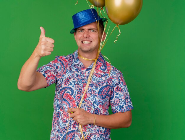 Chico de fiesta joven enojado con sombrero azul sosteniendo globos atados alrededor del cuello mostrando el pulgar hacia arriba aislado en verde