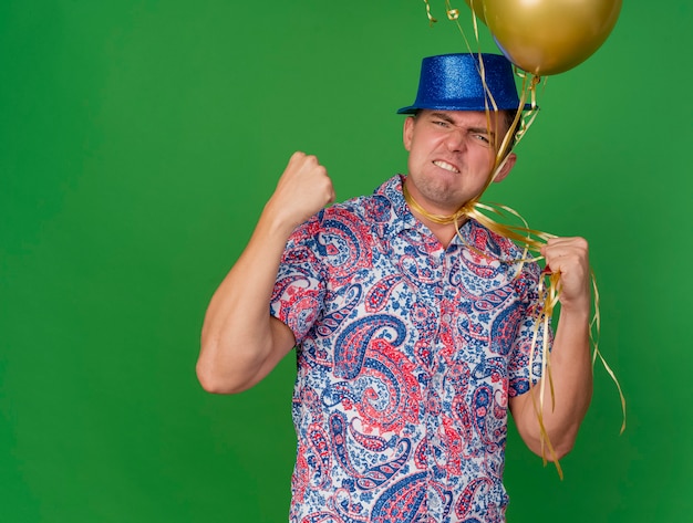 Chico de fiesta joven enojado con sombrero azul sosteniendo globos atados alrededor del cuello mostrando sí gesto aislado en verde