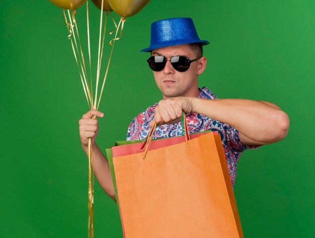 Chico de fiesta joven estricto con sombrero azul y gafas sosteniendo globos con bolsas de regalo aisladas en verde