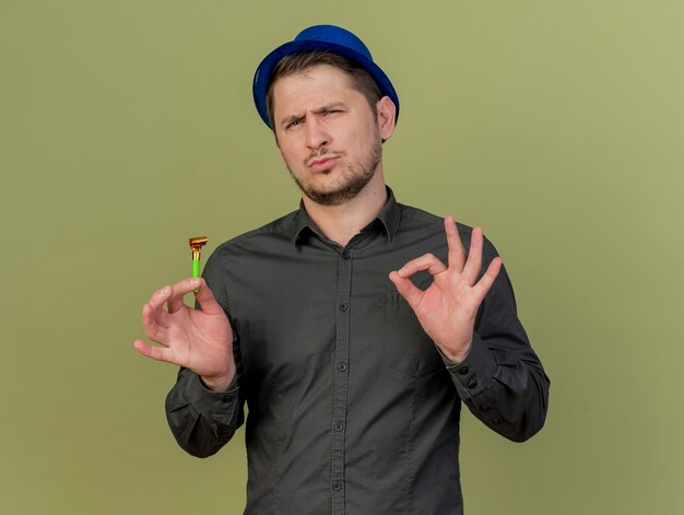 Chico de fiesta joven impresionado con camisa negra y sombrero azul sosteniendo el ventilador de fiesta mostrando gesto okey aislado en verde oliva