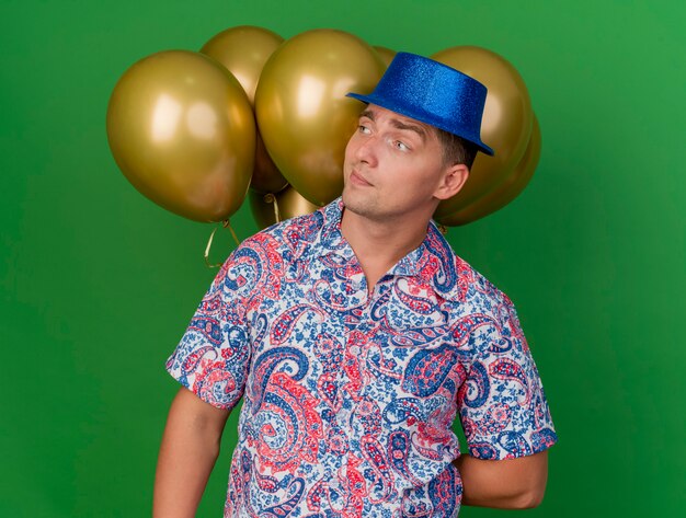 Chico de fiesta joven impresionado mirando al lado con sombrero azul de pie delante de globos aislados sobre fondo verde