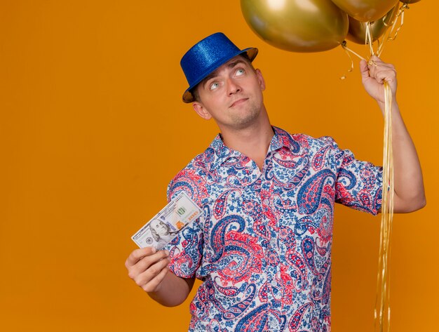 Chico de fiesta joven impresionado con sombrero azul sosteniendo globos con dinero en efectivo aislado en naranja