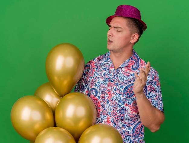 Chico de fiesta joven impresionado con sombrero rosa sosteniendo y mirando globos aislados en verde