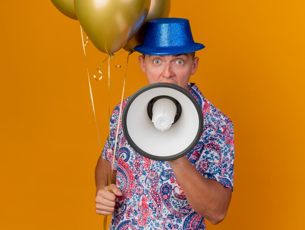 Chico de fiesta joven con sombrero azul sosteniendo globos y habla por altavoz aislado en naranja