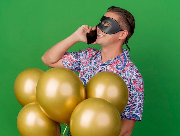 Foto gratuita chico de fiesta joven sonriente mirando a un lado con máscara de ojos de mascarada de pie entre globos y habla por teléfono aislado en verde