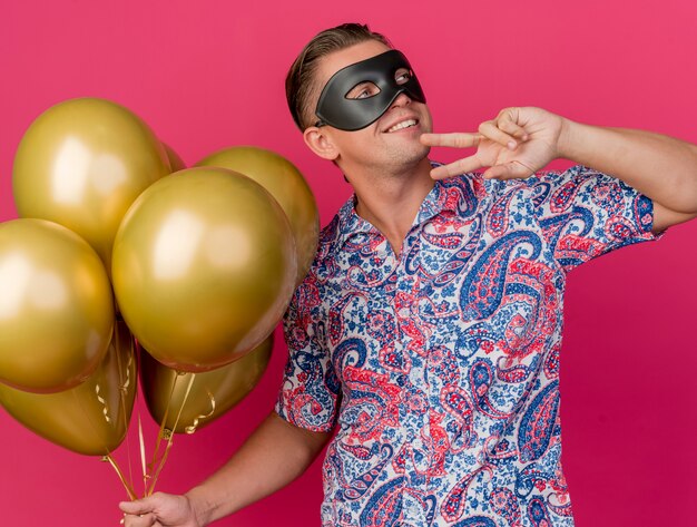 Foto gratuita chico de fiesta joven sonriente mirando a un lado con máscara de ojos de mascarada sosteniendo globos y mostrando gesto de paz aislado en rosa