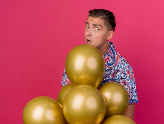 Chico de fiesta joven sorprendido con camisa colorida de pie detrás de globos aislados en rosa